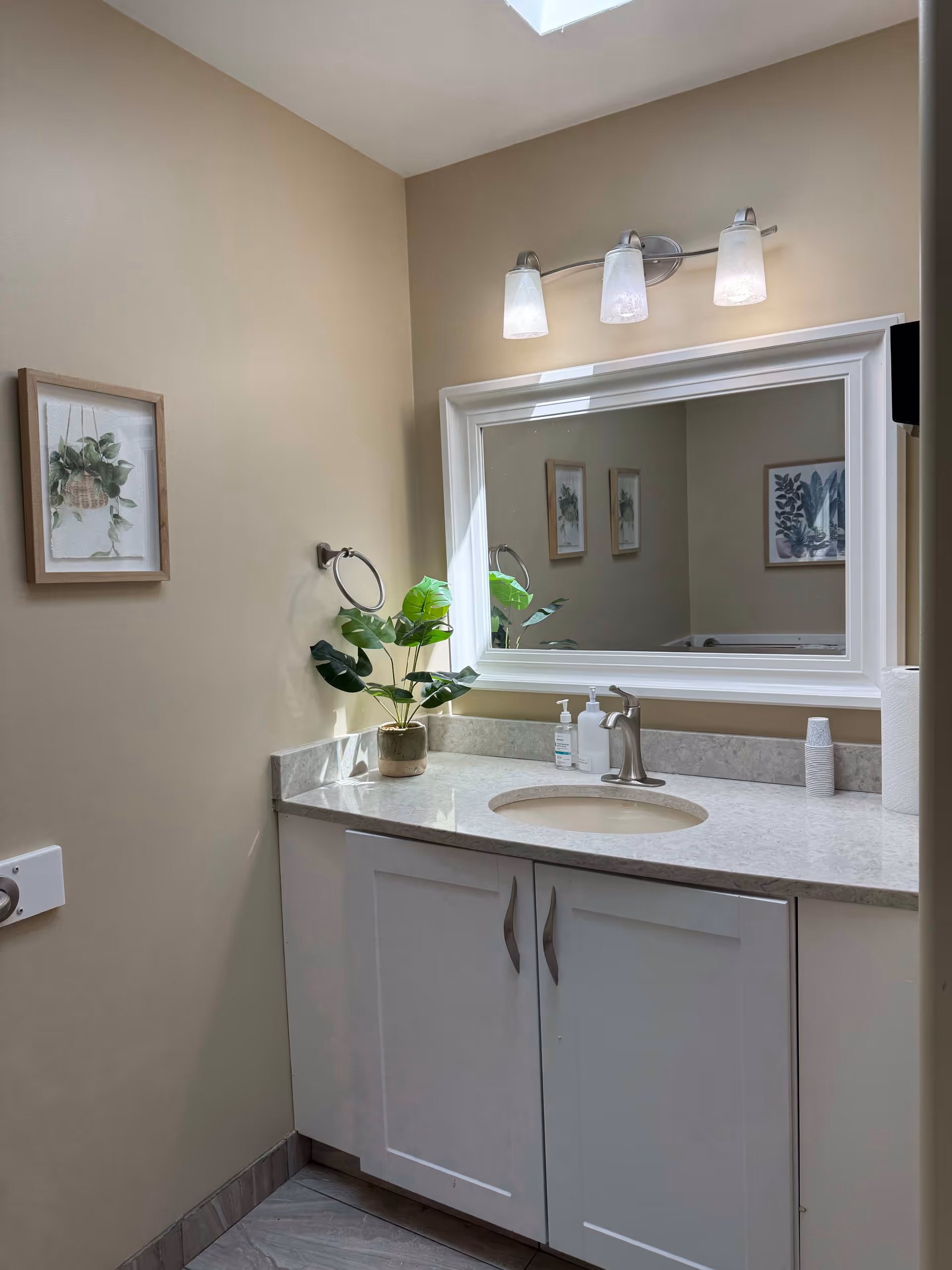 A bathroom vanity with a white countertop and sink, a silver faucet, and a large rectangular mirror above it. There are three light fixtures mounted above the mirror. On the countertop, there is a potted green plant, a soap dispenser, a bottle of hand sanitizer, and a roll of paper towels. The walls are beige, and there is a framed botanical print hanging on the left wall. The floor has gray tiles.