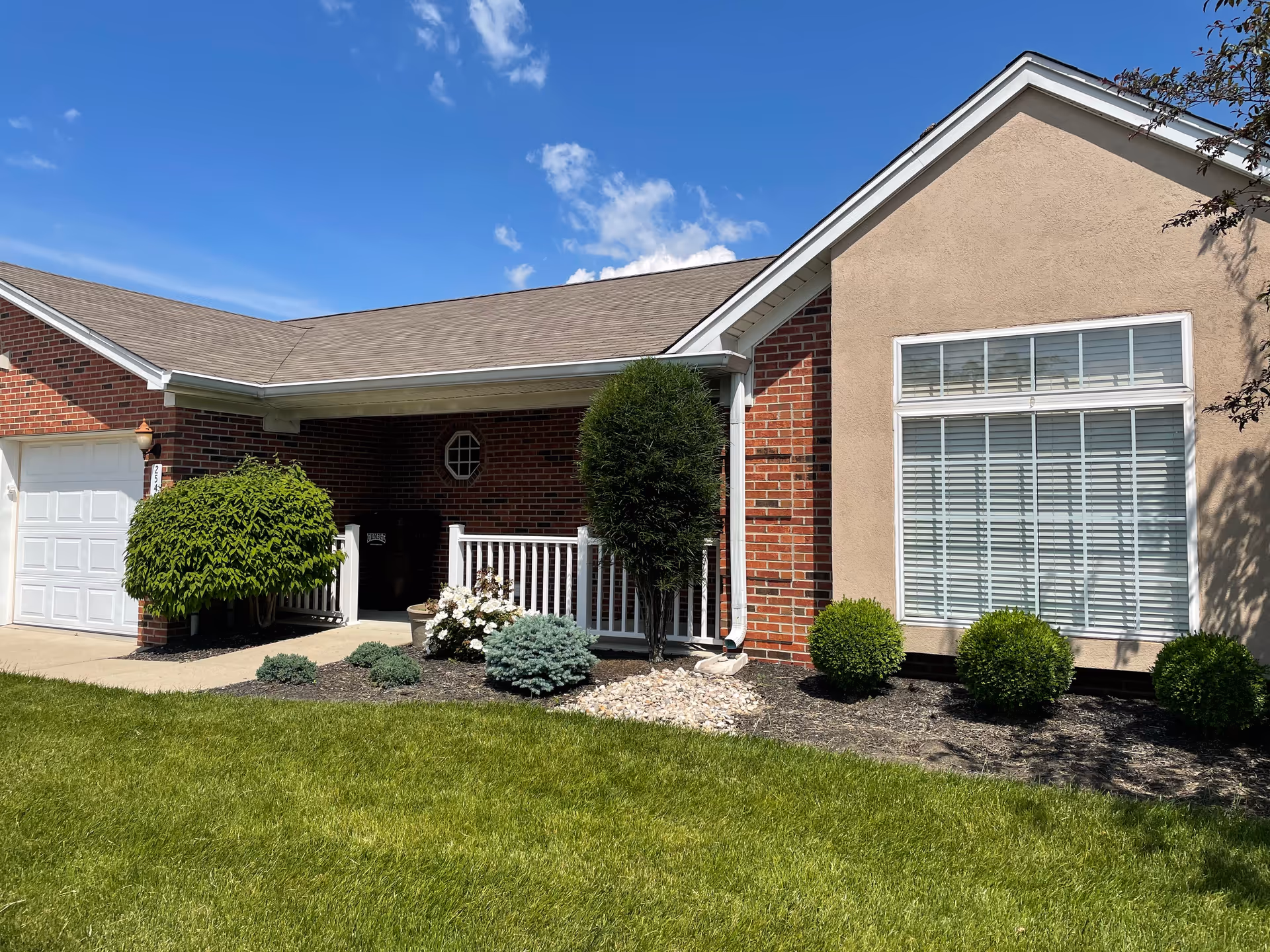Exterior view of a single-story residential building with a combination of red brick and beige stucco walls, a white garage door, a covered porch with white railing, and neatly trimmed bushes and landscaping in front under a blue sky with some clouds.