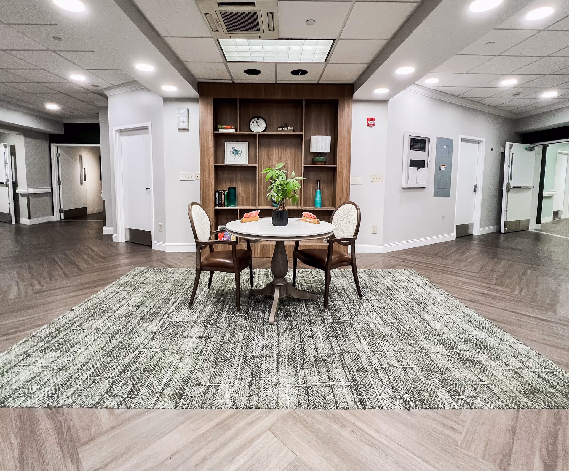 A well-lit interior common area with a round table and two chairs placed on a patterned rug. Behind the table is a wooden built-in shelving unit with decorative items including a clock, books, a lamp, and a plant. The space has light-colored walls, wood-patterned flooring, and multiple doors and hallways leading to other parts of the facility.