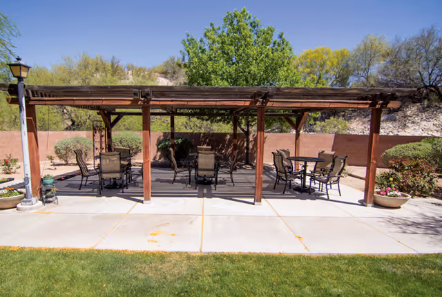 Outdoor patio area with a wooden pergola providing shade over several tables and chairs. The patio is surrounded by a low brick wall, desert plants, and trees under a clear blue sky.