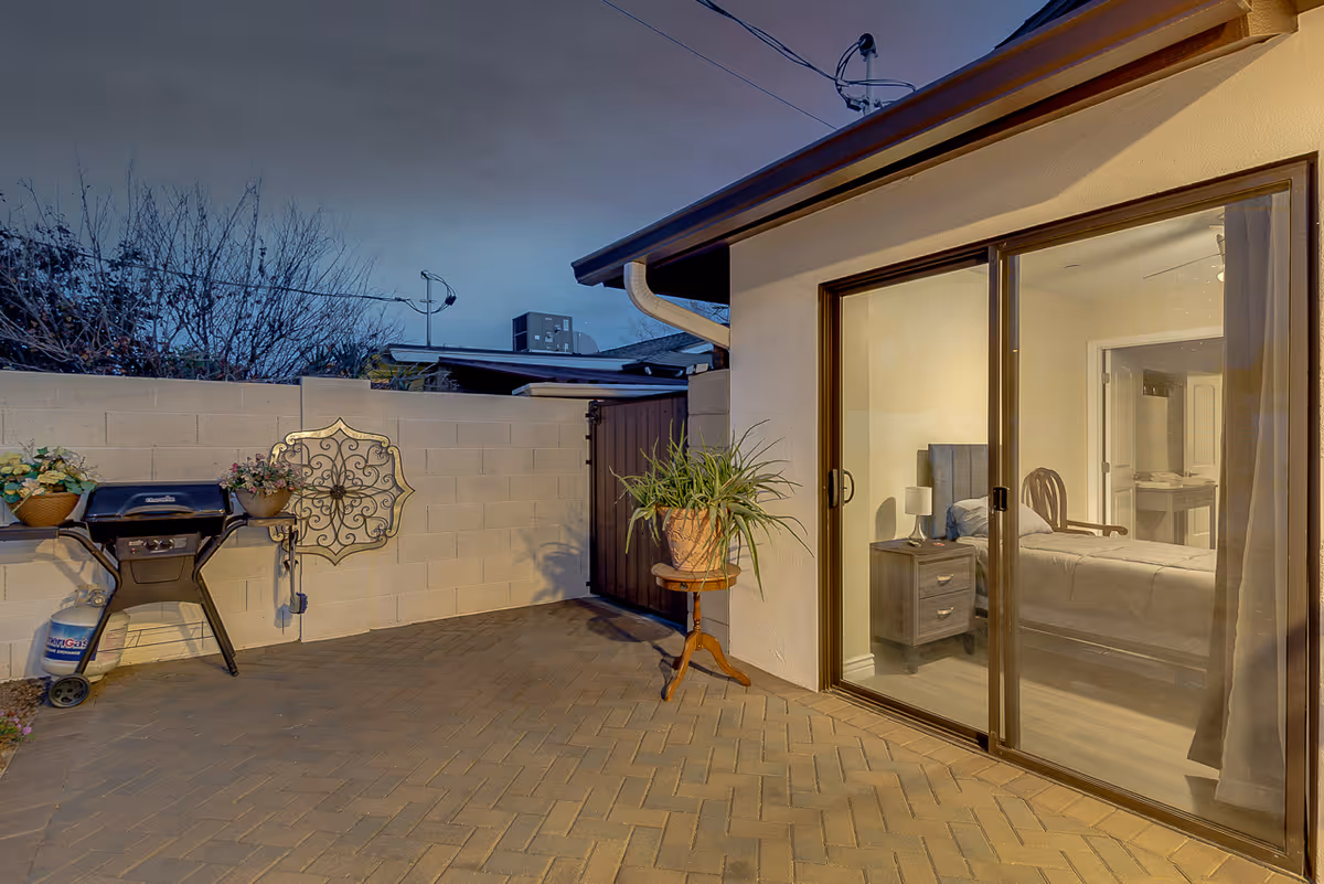 Outdoor patio area with a brick floor, a barbecue grill with a propane tank, potted plants, and a decorative wall hanging. A sliding glass door reveals a bedroom inside with a bed, nightstand, lamp, and chair.