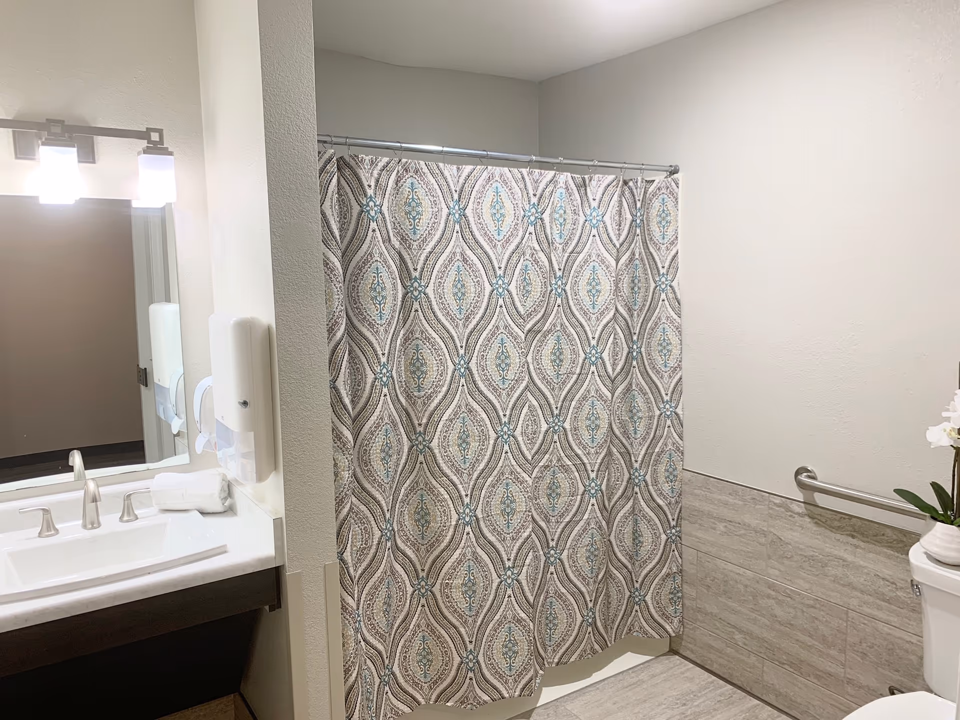 Bathroom with a patterned shower curtain, a white sink with silver faucet, a mirror with lights above it, a soap dispenser on the wall, a toilet with a white orchid plant on top, and a grab bar on the wall next to the toilet.