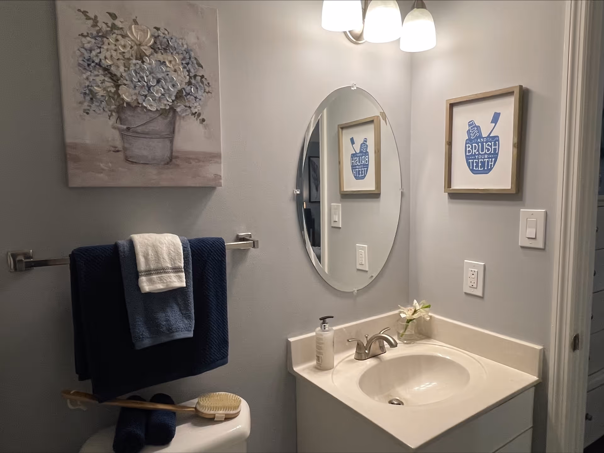 A bathroom with a white sink and countertop, an oval mirror above the sink, and a wall-mounted light fixture with three bulbs. There is a framed picture on the wall with the text 'Brush Your Teeth' and a toothbrush graphic. A towel rack holds two dark blue towels and one white towel. A hairbrush rests on top of a closed toilet lid. A floral painting hangs on the wall above the towel rack.