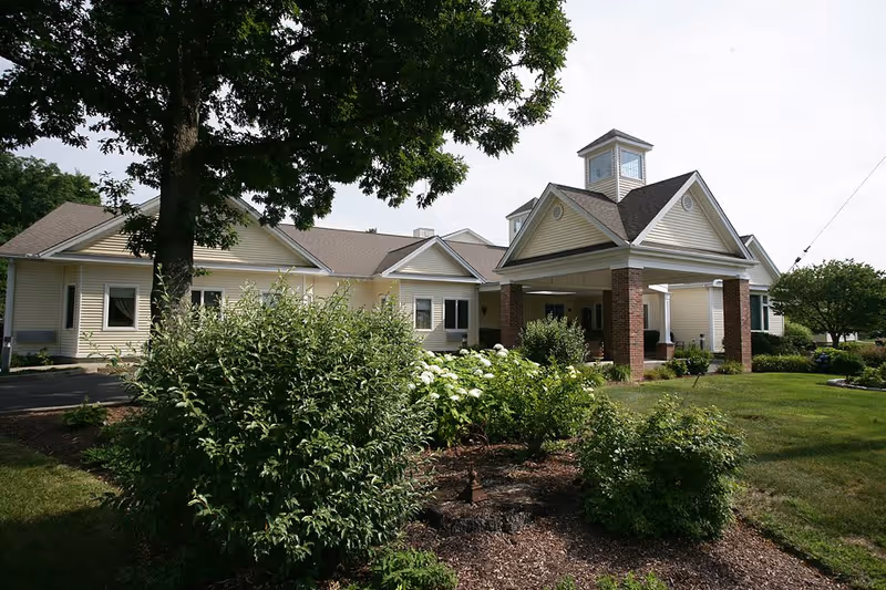 Exterior view of a single-story senior living facility building with beige siding, a covered entrance supported by brick columns, surrounded by green bushes, trees, and a well-maintained lawn under a partly cloudy sky.