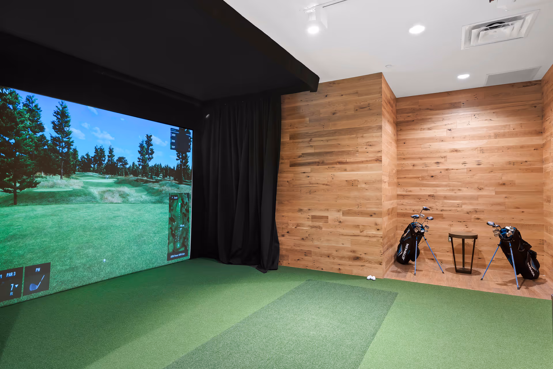 Indoor golf simulator room with a large screen displaying a golf course, green artificial turf flooring, wooden paneled walls, two golf bags with clubs, a small stool, and a few golf balls on the floor.