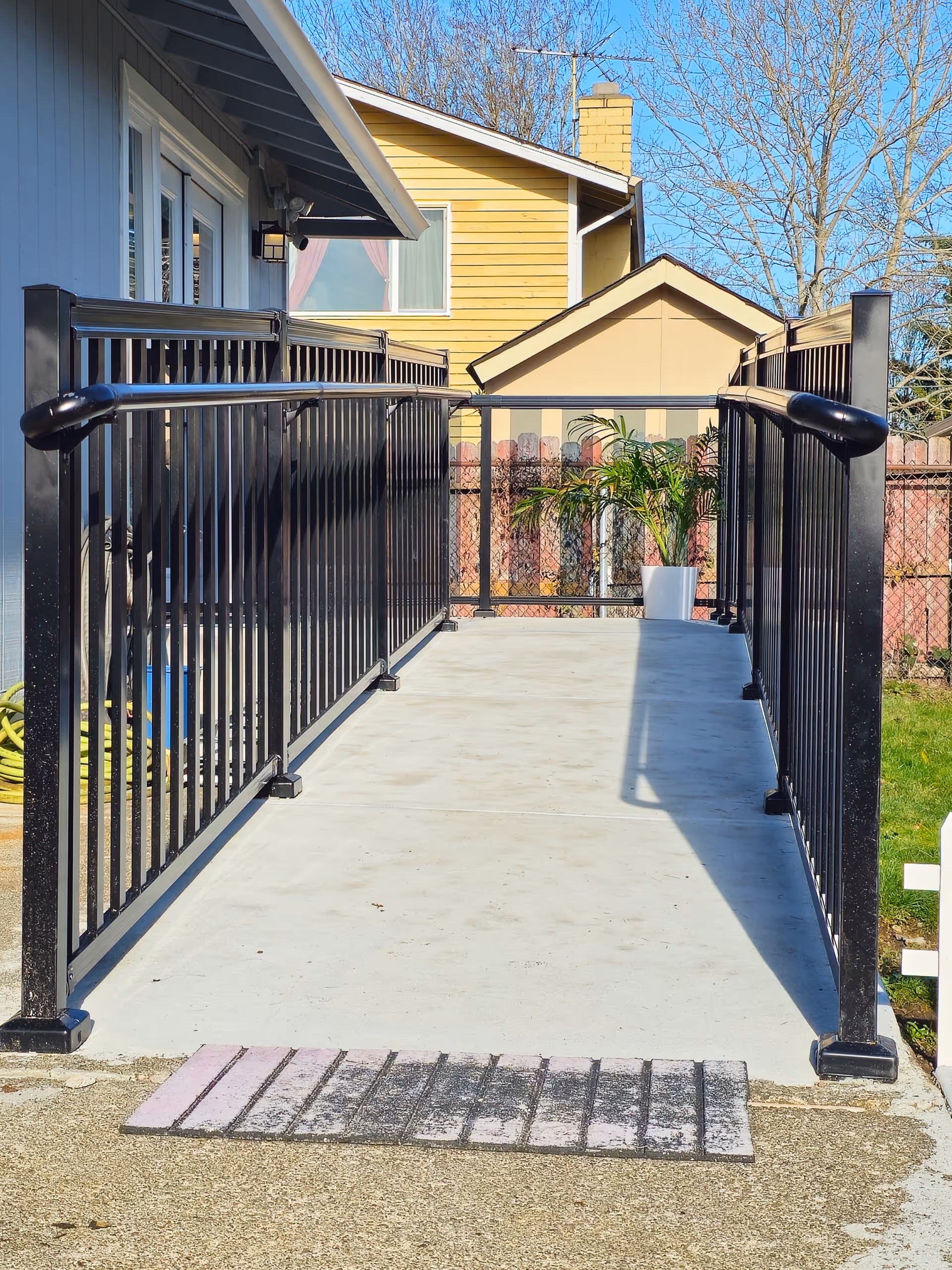 A concrete ramp with black railings leads to a doorway, surrounded by houses and greenery.