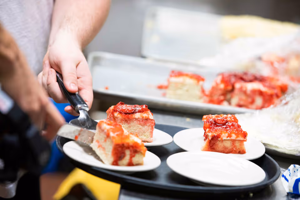 Close-up of a person serving slices of strawberry cake onto small white plates using a spatula in a kitchen or food preparation area.