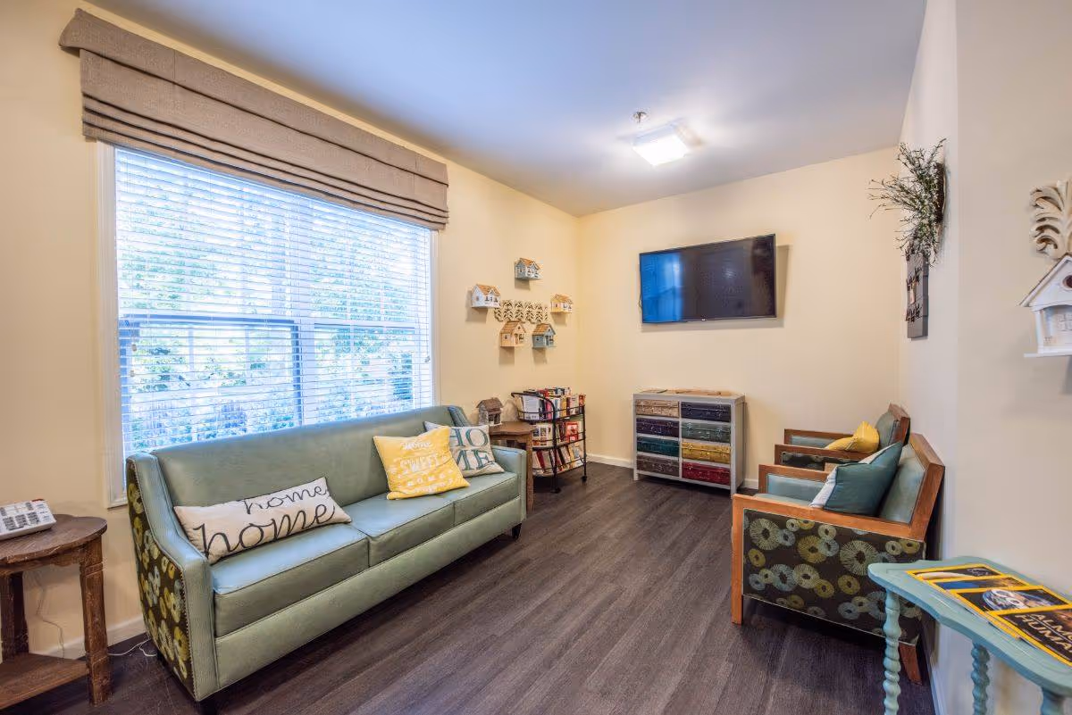 A cozy living room with a large window covered by blinds and a beige valance. There is a green sofa with decorative pillows, two armchairs with patterned sides and cushions, a small wooden side table, a colorful chest of drawers, a wall-mounted flat-screen TV, and wall decorations including small house-shaped shelves and a wreath.