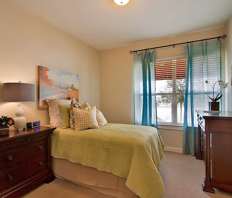A cozy bedroom with a single bed covered in a light green quilt and multiple decorative pillows. There is a wooden nightstand with a lamp, a digital clock, and a small plant. A large window with blue curtains lets in natural light, and a wooden dresser with a potted orchid is positioned near the window. A landscape painting hangs above the bed.