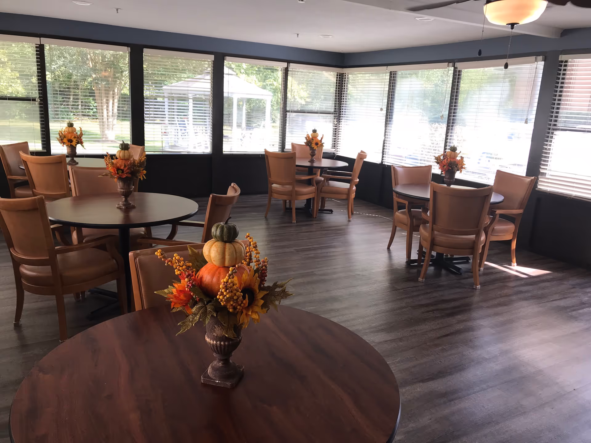 Sunlit dining room with round wooden tables, chairs, and fall-themed centerpieces in front of large windows.