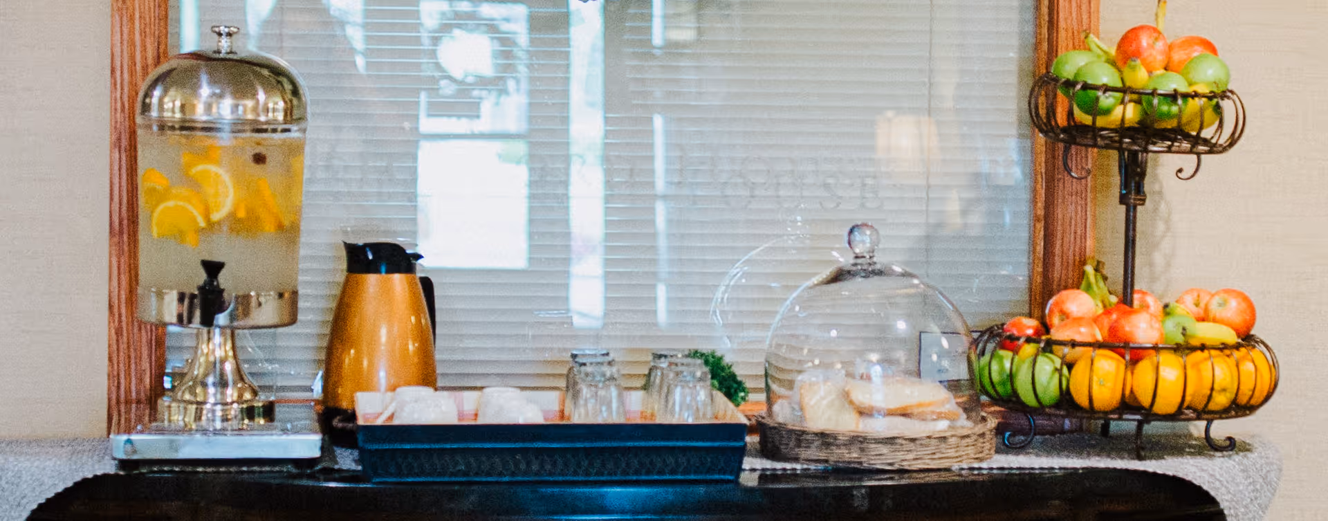A refreshment station with a glass dispenser filled with water and lemon slices, a thermal coffee carafe, a tray holding cups and glasses, and a two-tiered fruit basket containing apples, bananas, and pears, set against a window with blinds.