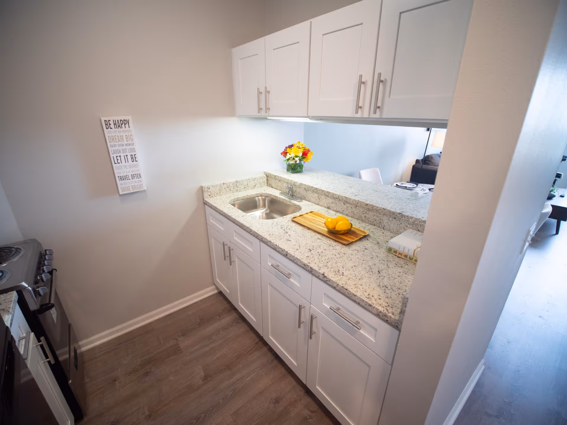 A small kitchen area with white cabinets, a granite countertop, a stainless steel sink, and a stove. There is a wooden cutting board with lemons on the counter and a vase of colorful flowers near the pass-through window that opens to a living room area. A decorative sign with inspirational quotes is mounted on the wall.