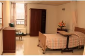 A simple, clean bedroom in a senior living facility featuring a hospital-style bed with a patterned bedspread, a wooden wardrobe, a small table with a flower arrangement, a chair, and a window with curtains allowing natural light.