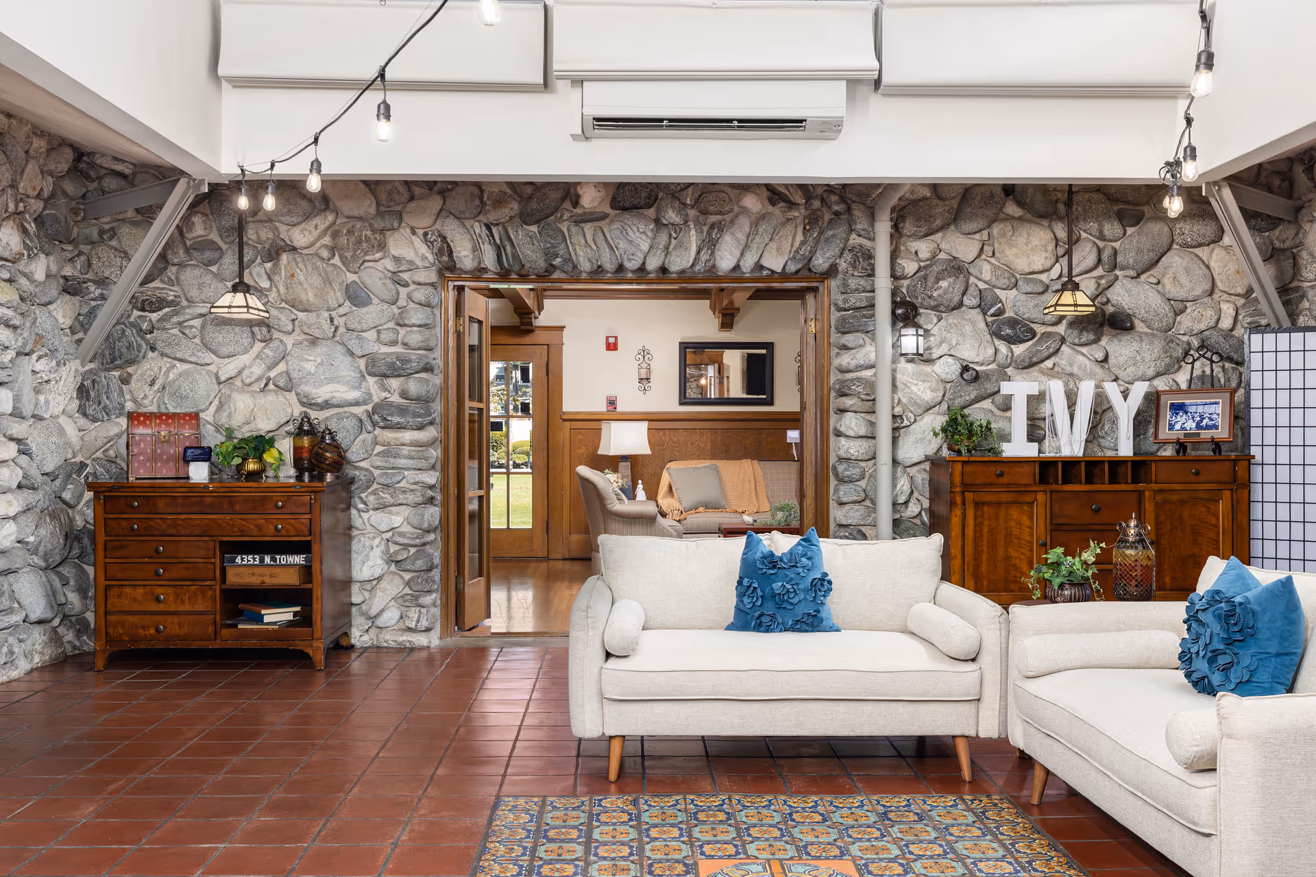 A cozy living room area with stone walls and terracotta tile flooring. The room features two beige sofas with blue decorative pillows, a colorful patterned rug, and wooden cabinets against the stone walls. One cabinet has large white letters spelling 'IVY' along with plants and framed photos. Through an open doorway, another seating area with a lamp and wooden paneling is visible.