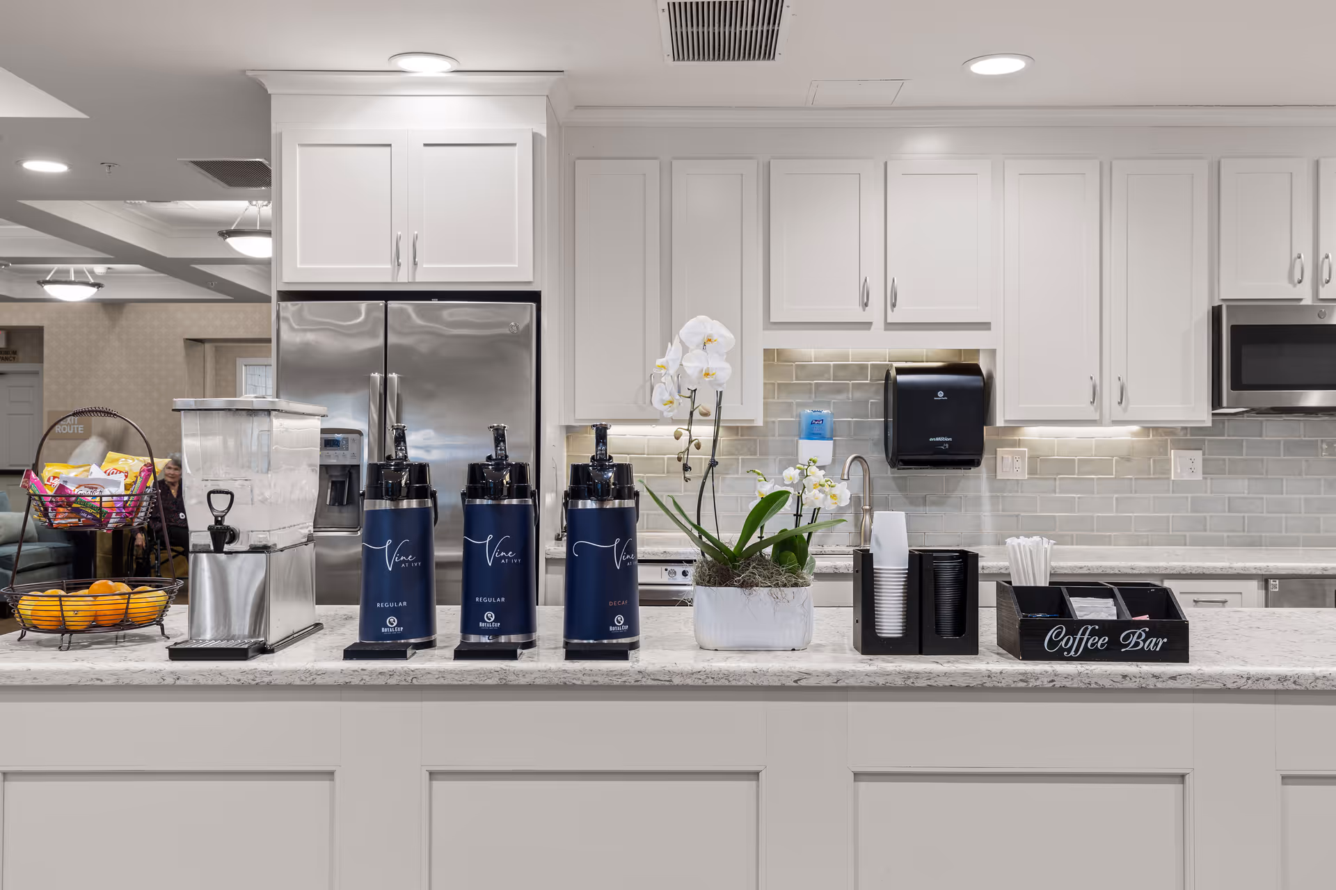 A modern coffee bar area in a senior living facility kitchen with white cabinets, a stainless steel refrigerator, coffee dispensers labeled Vine at Ivy, a water dispenser, a basket of snacks and oranges, a white orchid plant, and various coffee supplies including cups, stirrers, and sweeteners.
