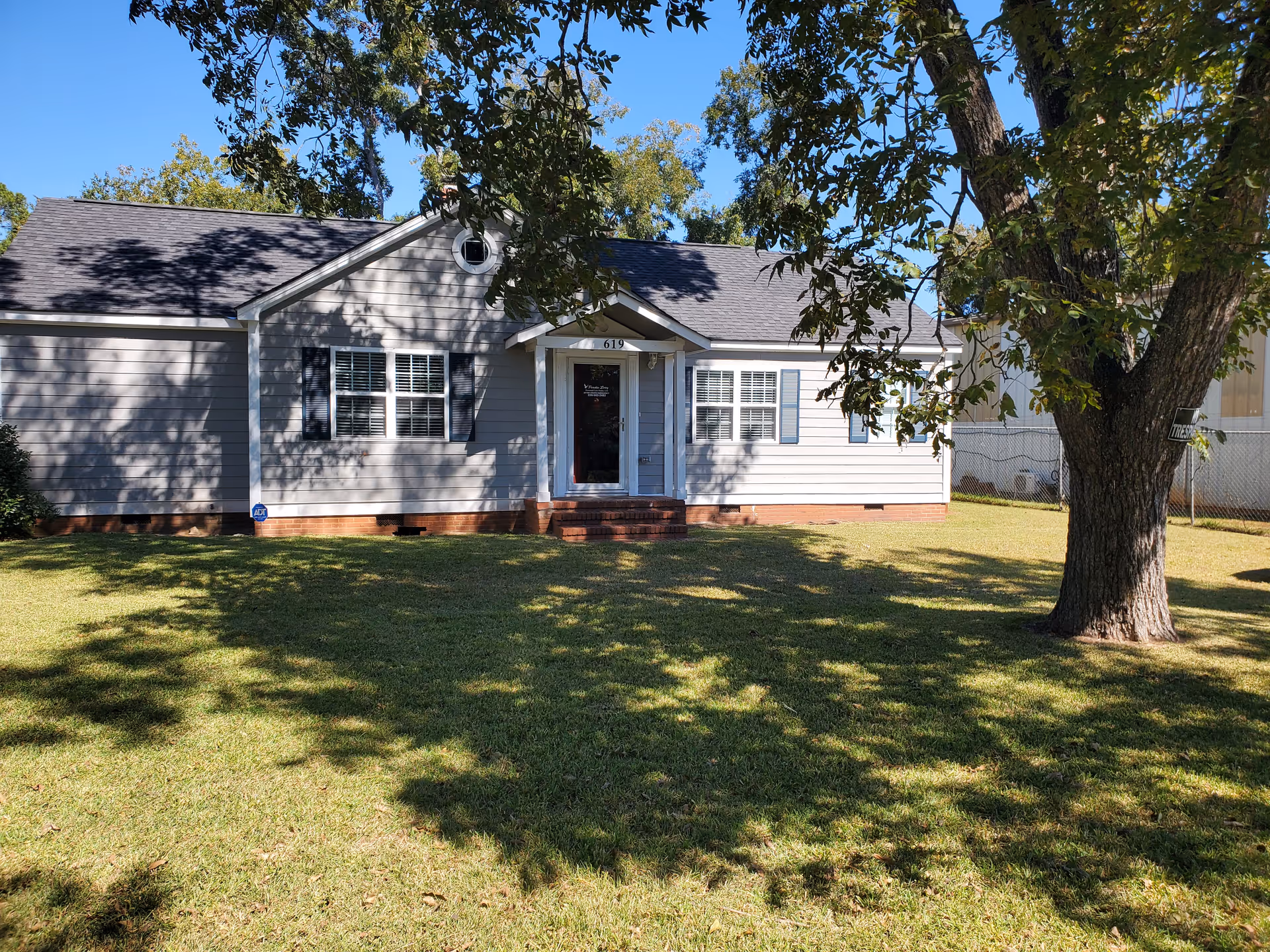 Front exterior view of a single-story house with gray siding, white trim, and black shutters. There is a small porch with steps leading to a glass front door. The house is surrounded by a well-maintained lawn and a large tree casting shadows on the grass. The sky is clear and blue.