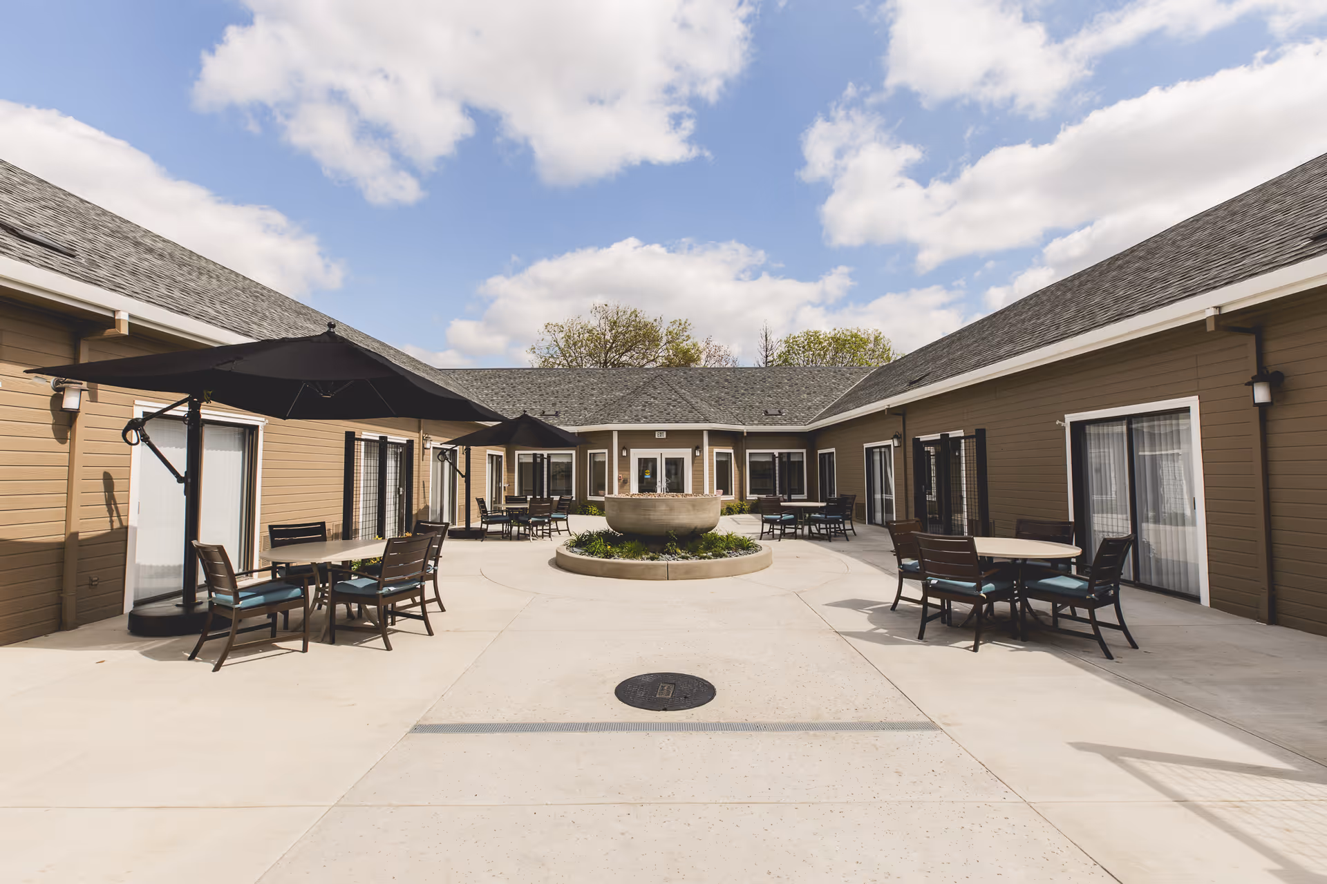 Outdoor courtyard area at Orangeburg Manor with several tables and chairs under large black umbrellas, surrounded by single-story brown buildings with white trim and sliding glass doors, under a partly cloudy sky.