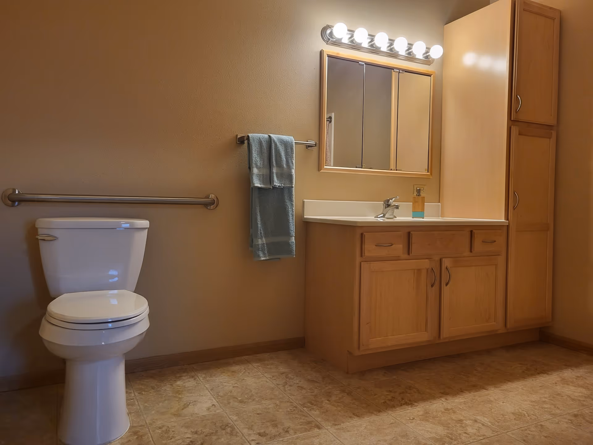 A bathroom with a white toilet, a towel rack with a blue towel, a wooden vanity with a sink, a soap dispenser, a large mirror with lights above it, and a tall wooden cabinet. The floor is tiled and the walls are painted beige.