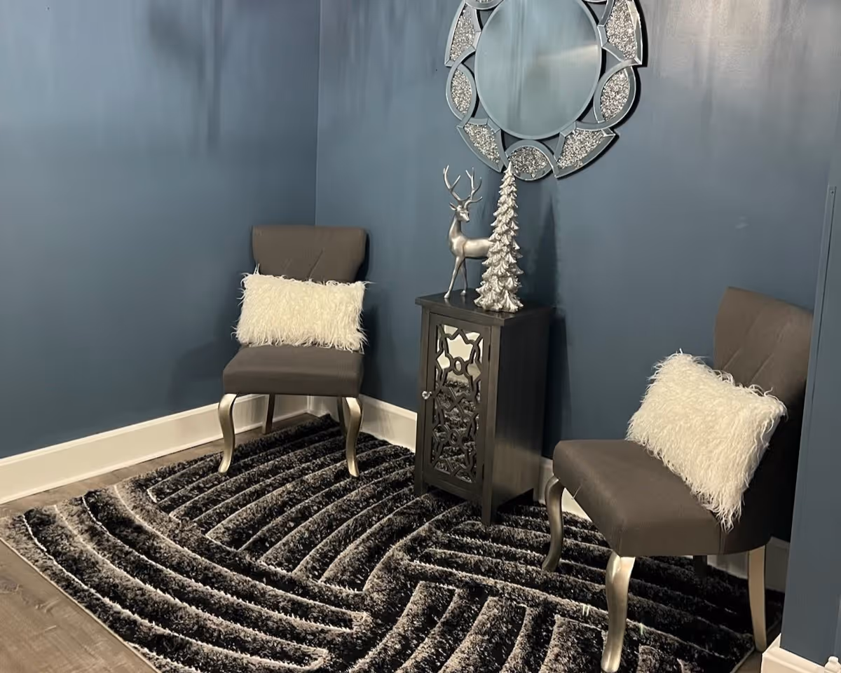Two upholstered chairs with white faux-fur pillows flank a decorative cabinet under a round mirror in a cozy blue-walled seating nook.