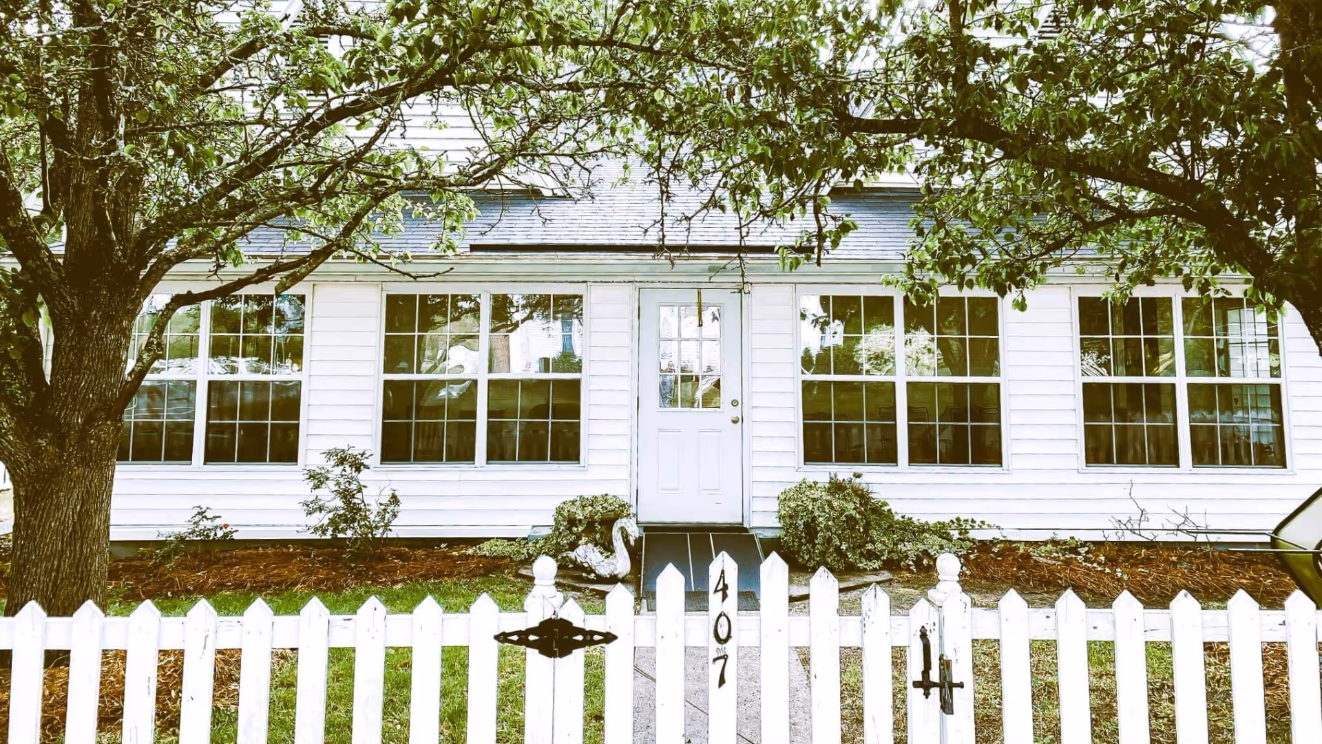 Front exterior of a white house with large windows, a centered door, trees and a white picket fence numbered 407.