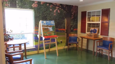 A cozy interior room with green flooring and a large window letting in natural light. The walls feature a mural of a garden with trees and flowers. There is an easel with a painting of a landscape, a small round table with chairs, and a corner table with two blue cushioned chairs. The wall on the right has a window with red shutters and a decorative item on the table beneath it.