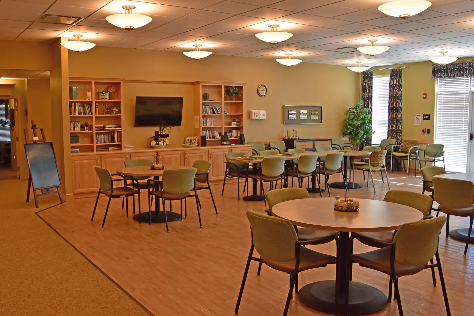 A spacious common area in an assisted living facility with multiple round tables and green cushioned chairs arranged for group seating. The room features a built-in wooden bookshelf with books and decorative items, a wall-mounted flat-screen TV, and large windows with patterned curtains allowing natural light to enter. The flooring is a combination of carpet and wood, and the ceiling has multiple light fixtures.
