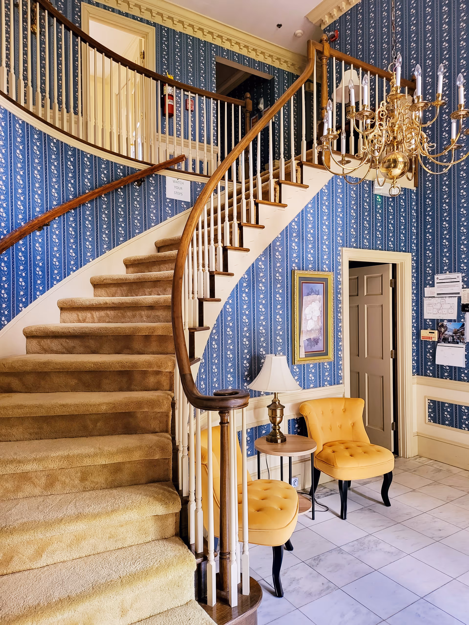 Interior view of a senior living facility featuring a curved staircase with beige carpet and wooden handrails. The walls are covered with blue floral wallpaper. Two yellow upholstered chairs and a small round table with a lamp are placed near the staircase. A gold chandelier hangs from the ceiling, and there is a framed picture on the wall next to an open doorway.