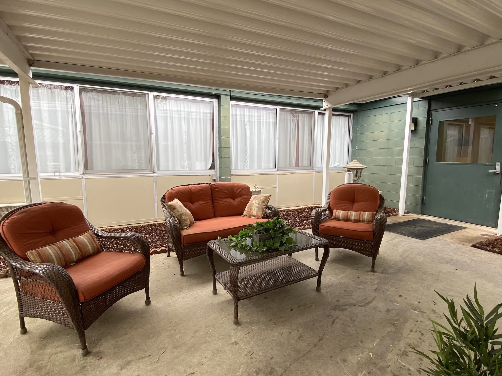 Covered outdoor seating area with three wicker chairs and a wicker loveseat, all with rust-colored cushions and decorative pillows. A glass-top wicker coffee table with a green plant centerpiece is in the middle. The area is enclosed with windows covered by white curtains and has a green door on the right side.