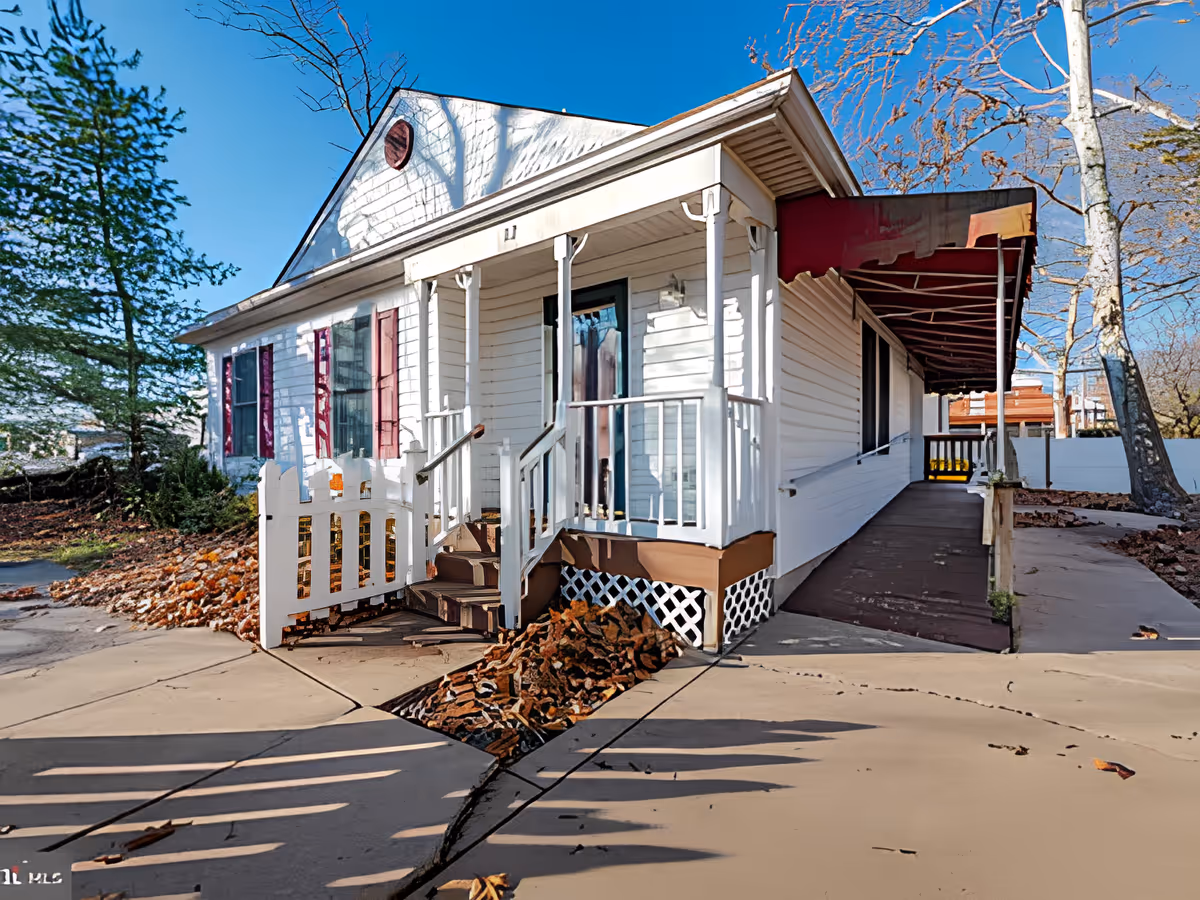 Exterior view of a white single-story building with a small porch, steps, and a wheelchair ramp. The building has red shutters and a red awning over the ramp. There are trees and fallen leaves around the building under a clear blue sky.