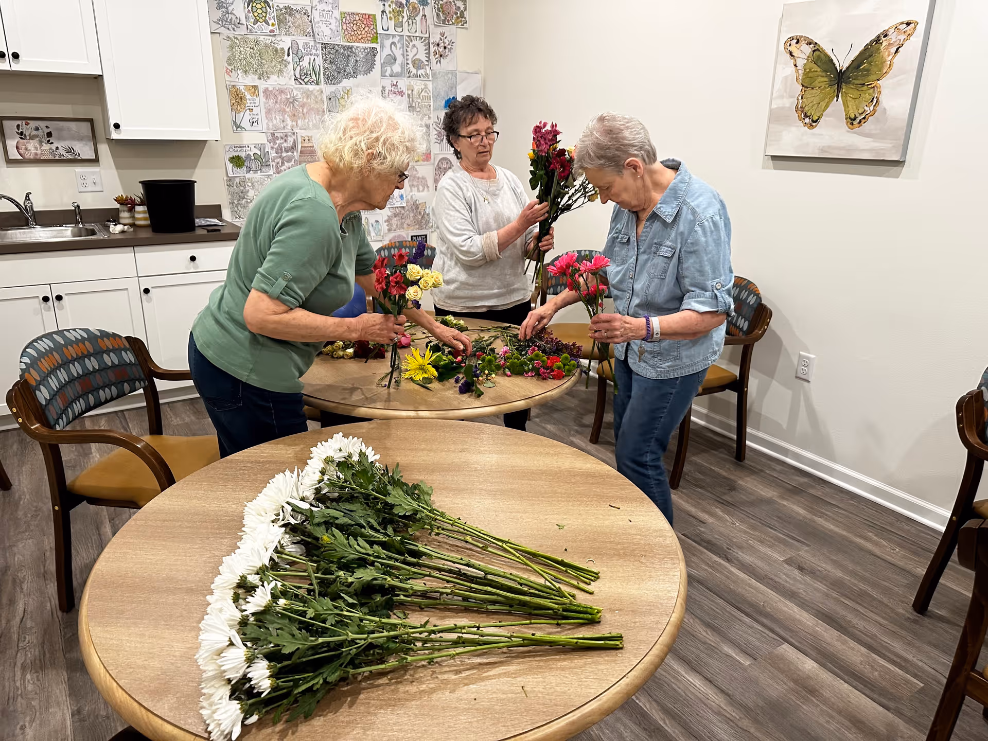 Three elderly women arranging colorful flowers on a round wooden table in a room with wooden flooring, white walls, and a butterfly painting. The room has cabinets and a sink in the background, and a wall decorated with various floral and inspirational drawings.