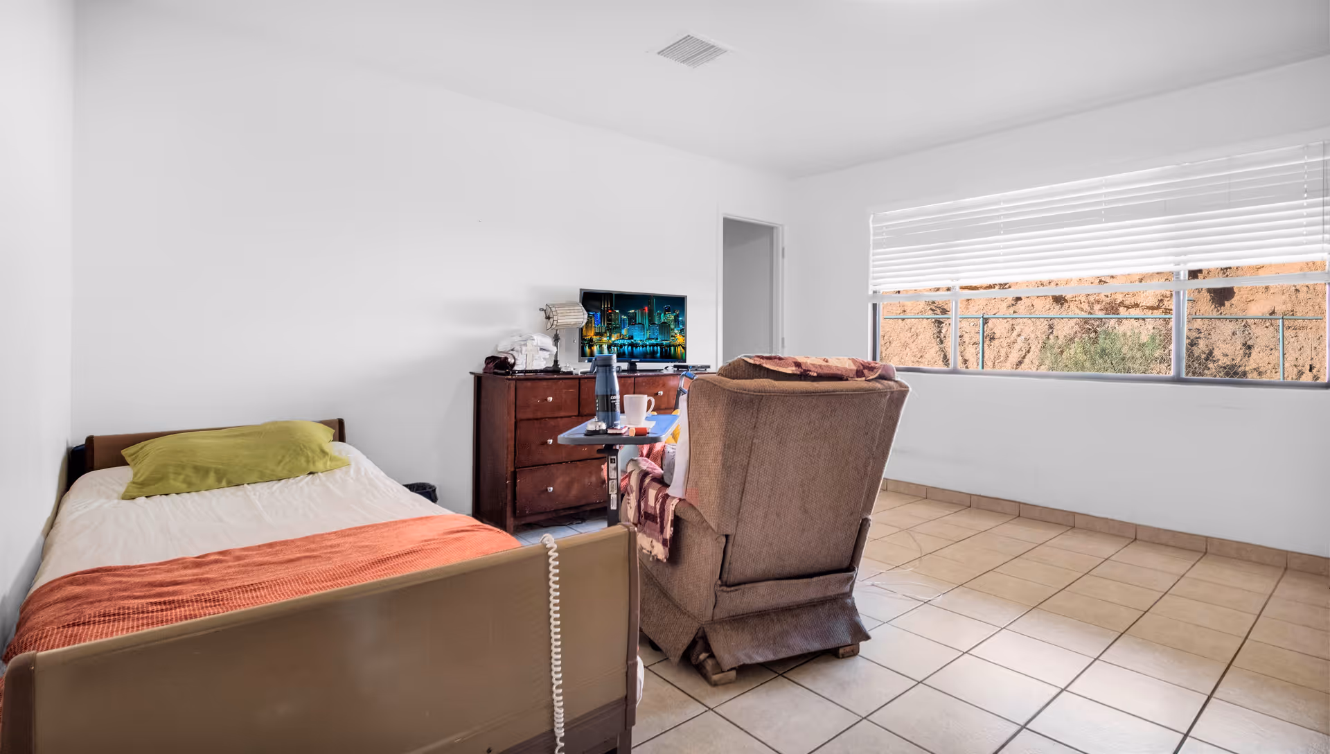 A simple bedroom with a single bed with a green pillow and orange blanket, a brown recliner chair with a blanket draped over it, a wooden dresser with a TV on top, and a large window with white blinds showing an outdoor view of rocky terrain.