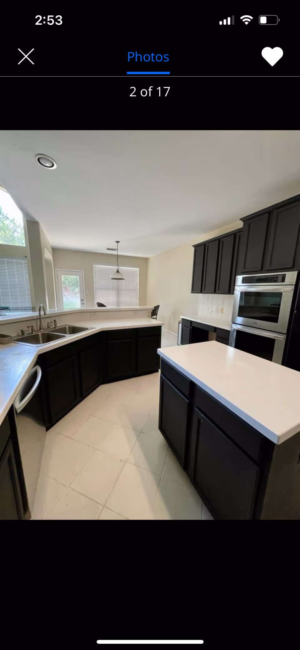 A modern kitchen with black cabinets and white countertops. The kitchen features a double sink, a built-in stainless steel oven, and a small island in the center. There is a window with blinds and a hanging light fixture above the dining area in the background.