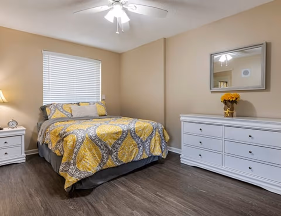 A bedroom with a bed covered in a yellow and gray patterned comforter, a white nightstand with a lamp and clock, a white dresser with a vase of yellow flowers, a mirror above the dresser, and a ceiling fan with lights.