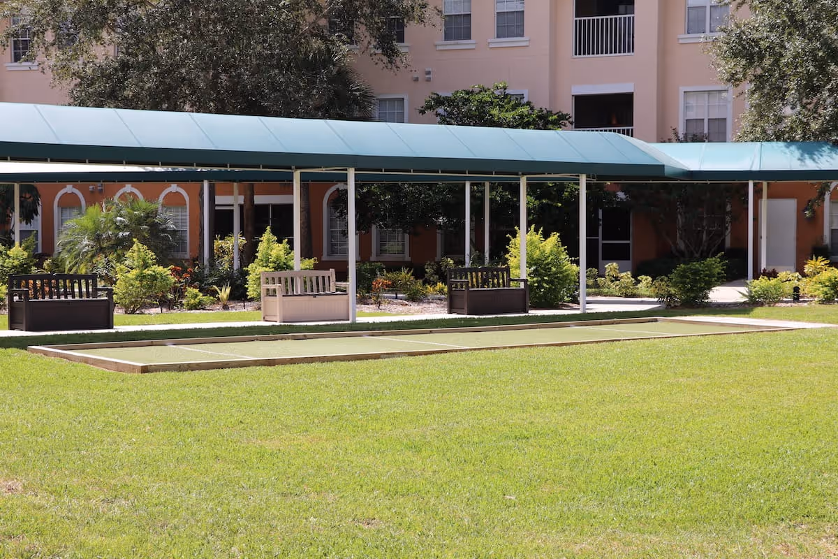 Outdoor area at Aston Gardens At Pelican Pointe featuring a covered walkway with benches underneath, surrounded by green grass, shrubs, and trees, with a multi-story building in the background.