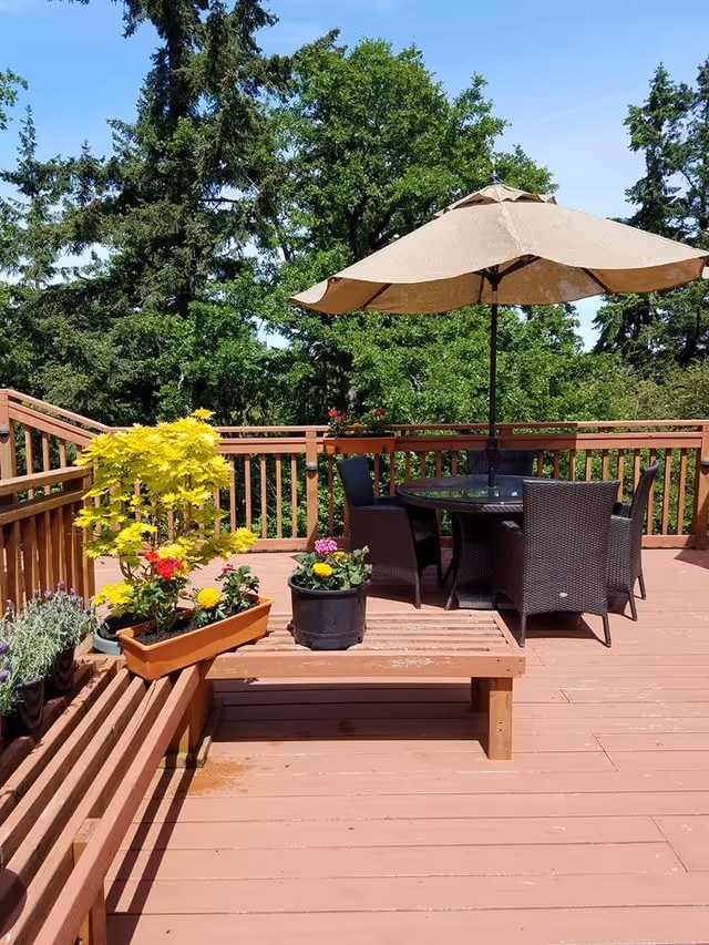 Outdoor wooden deck area with a round glass table and four wicker chairs under a large beige patio umbrella. Several potted plants with colorful flowers are placed on the deck railing and a wooden bench. Tall green trees and a clear blue sky are visible in the background.