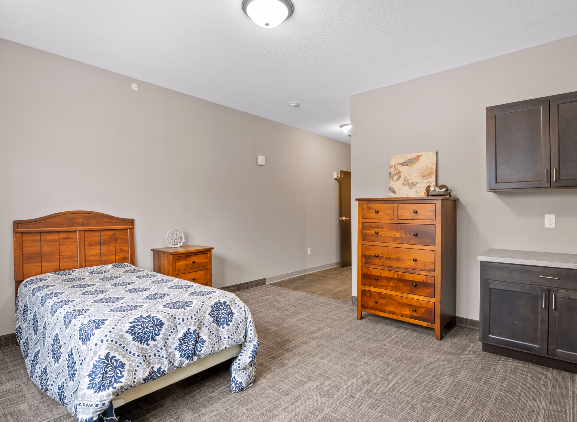 Furnished bedroom with a single bed, wooden nightstand and tall dresser against neutral walls and carpeted floor.