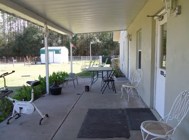Covered outdoor patio area with several metal chairs, a table, exercise bike, and a view of a fenced yard with trees and a small white shed in the background.