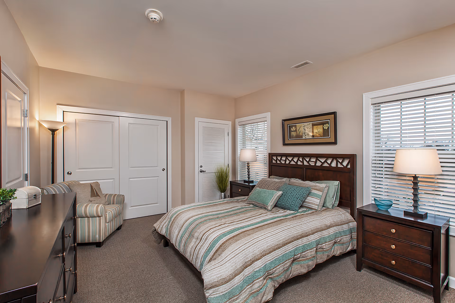 A neatly arranged bedroom with a double bed featuring striped bedding in shades of green, beige, and brown. The room includes two dark wooden nightstands with lamps, a framed artwork above the bed, a striped armchair, a dresser, and two windows with blinds letting in natural light. The walls are painted beige and the floor is carpeted.