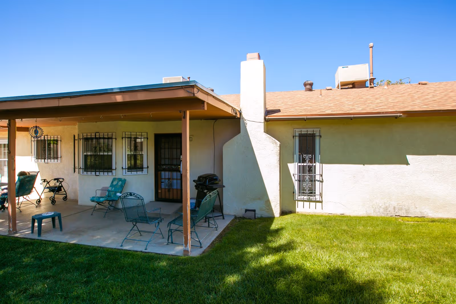 Outdoor patio area of a single-story building with a covered seating area. The patio has metal chairs with cushions, a small table, and a barbecue grill. The building has barred windows and a door, with a well-maintained green lawn in the foreground under a clear blue sky.