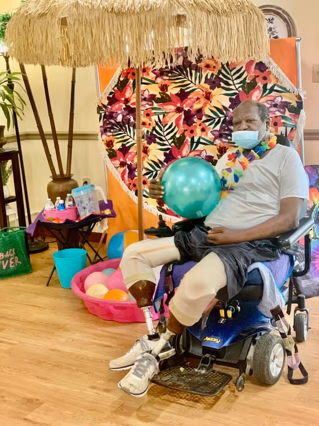 A man with prosthetic legs sitting in a motorized wheelchair indoors, holding a teal balloon. He is wearing a face mask and a colorful lei around his neck. Behind him is a vibrant floral backdrop with an orange border, a thatched umbrella, and a small table with water bottles and other items. There is also a pink plastic tub filled with colorful balloons on the floor.