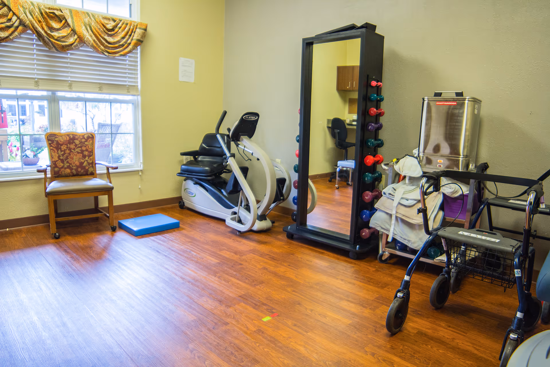 Small fitness/therapy room with a recumbent exercise bike, mirror with colorful dumbbells, a walker, a chair by a window, and wood flooring.