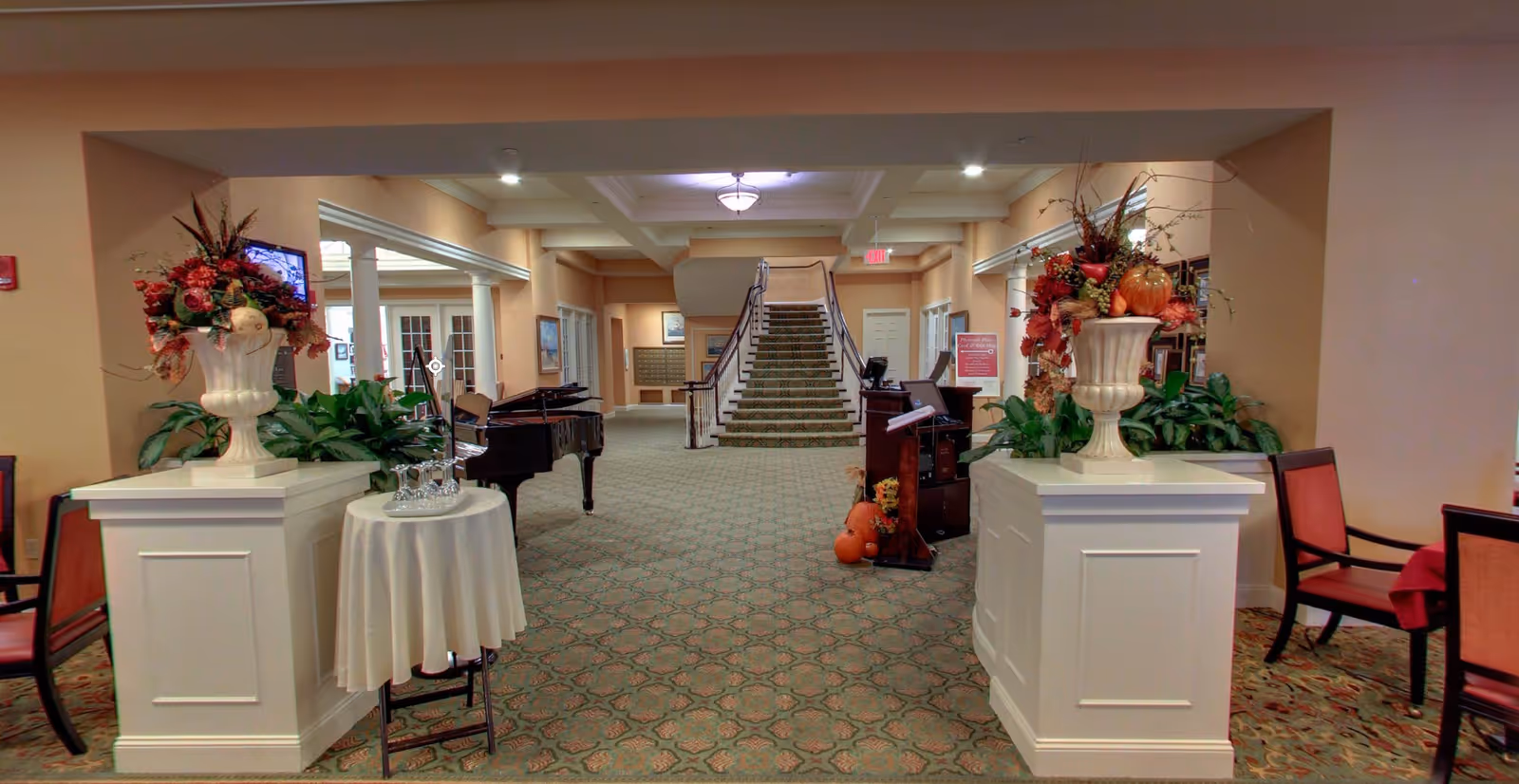 A decorated senior living facility lobby with a grand piano, floral arrangements, seating, and a central staircase.