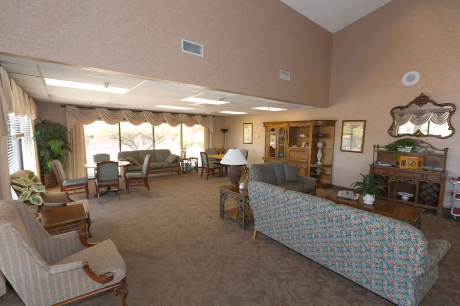 A spacious lounge with upholstered sofas and armchairs, tables, cabinets, and large windows letting in natural light.