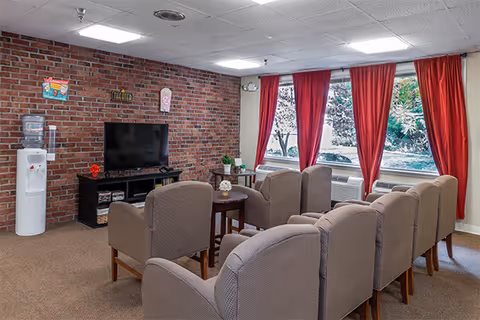 A cozy common room with a brick accent wall featuring a flat-screen TV on a black stand. There are eight beige upholstered chairs arranged in two rows facing the TV. A small round wooden table with a flower arrangement is placed between the chairs. Large windows with red curtains allow natural light to enter the room. A white water cooler is positioned against the brick wall.