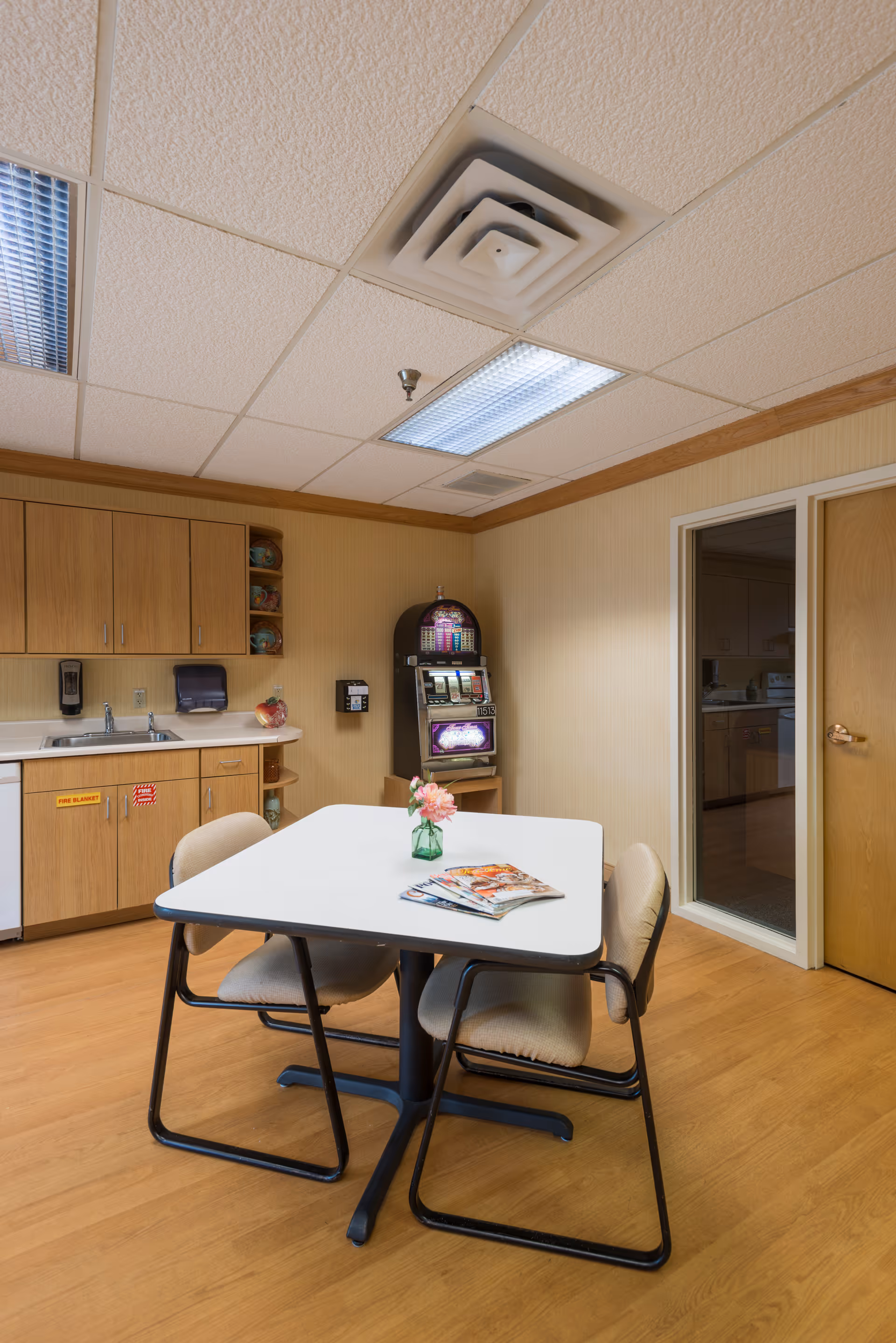 A small room with a white table and four beige chairs around it. On the table, there is a small green vase with pink flowers and some magazines. The room has wooden flooring and light-colored walls with wooden cabinets and a sink on one side. A slot machine is placed against the far wall. There is a door with a glass panel leading to another room.