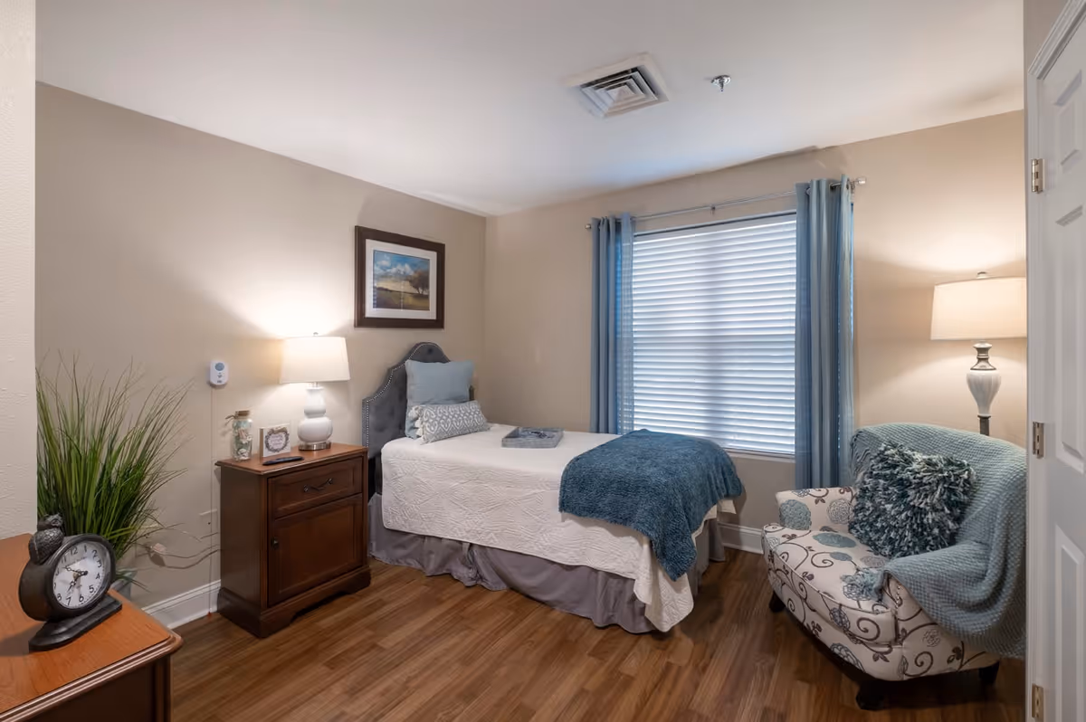 A cozy bedroom with a single bed covered in white bedding and a blue throw blanket. There is a wooden nightstand with a lamp, a clock, and decorative items next to the bed. A patterned armchair with a blue throw and pillow is placed near a window with blue curtains. The room has wooden flooring and neutral-colored walls with a framed picture hanging above the bed.
