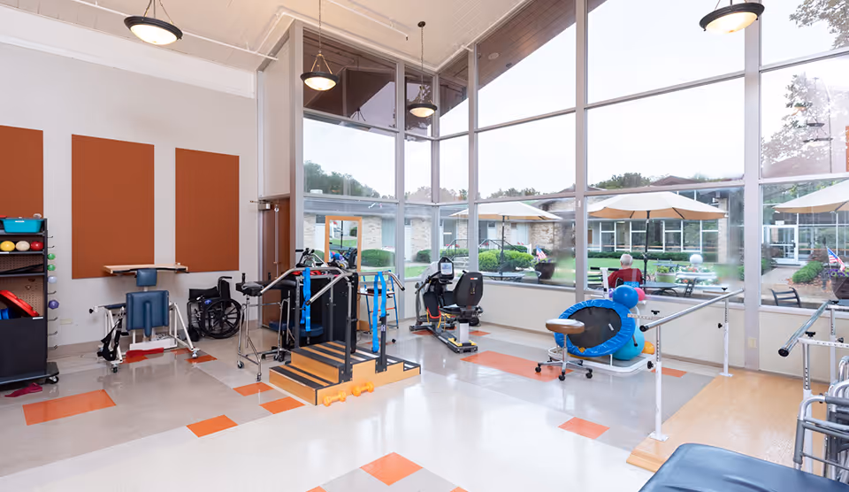 A bright rehabilitation room with large floor-to-ceiling windows overlooking an outdoor courtyard. The room contains various physical therapy equipment including parallel bars, exercise bikes, a wheelchair, therapy balls, and other rehabilitation tools. The floor has a pattern of orange and beige tiles, and the walls have three large orange acoustic panels. Outside, a person is sitting under a large umbrella in the courtyard.