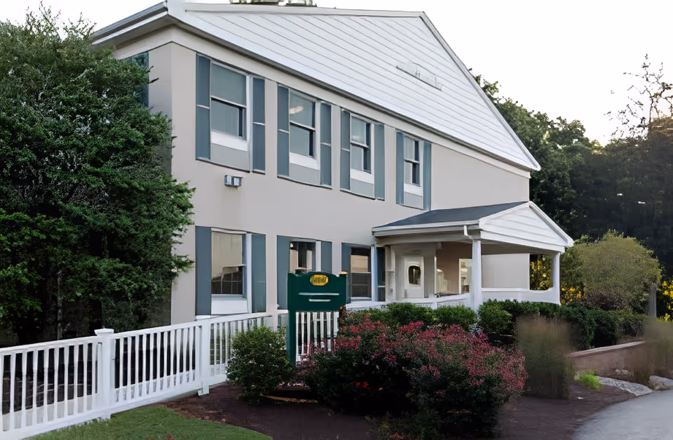 Exterior view of a two-story building with white siding and blue shutters, surrounded by greenery and bushes. There is a white railing along a walkway leading to a covered porch entrance.