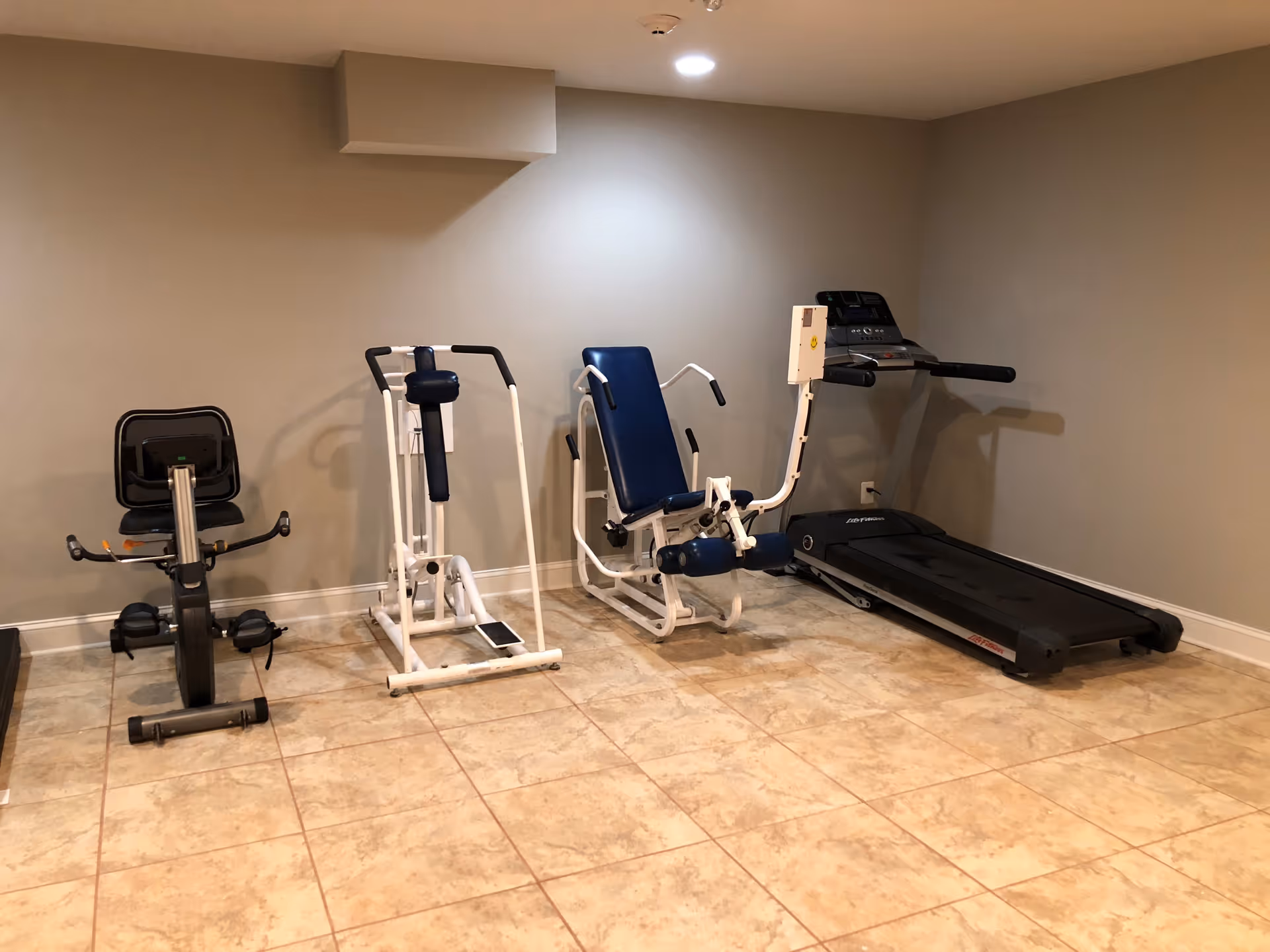 A small exercise room with beige tiled floor and light gray walls, containing a recumbent exercise bike, two white and blue strength training machines, and a black treadmill.