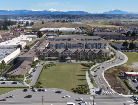 Aerial view of the assisted living and memory care building with a large front lawn, parking areas, surrounding roads, and mountains in the background.